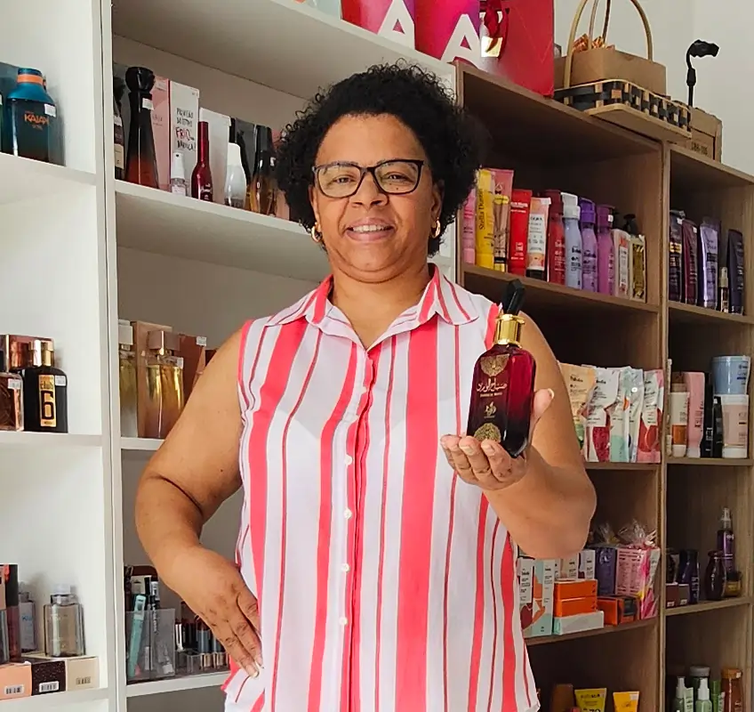Proprietária da Val Cosméticos e Presentes em Londrina, segurando na mão um frasco de perfume árabe importado e original sorrindo, dentro da loja, com as prateleiras de perfumes e cosméticos ao fundo.