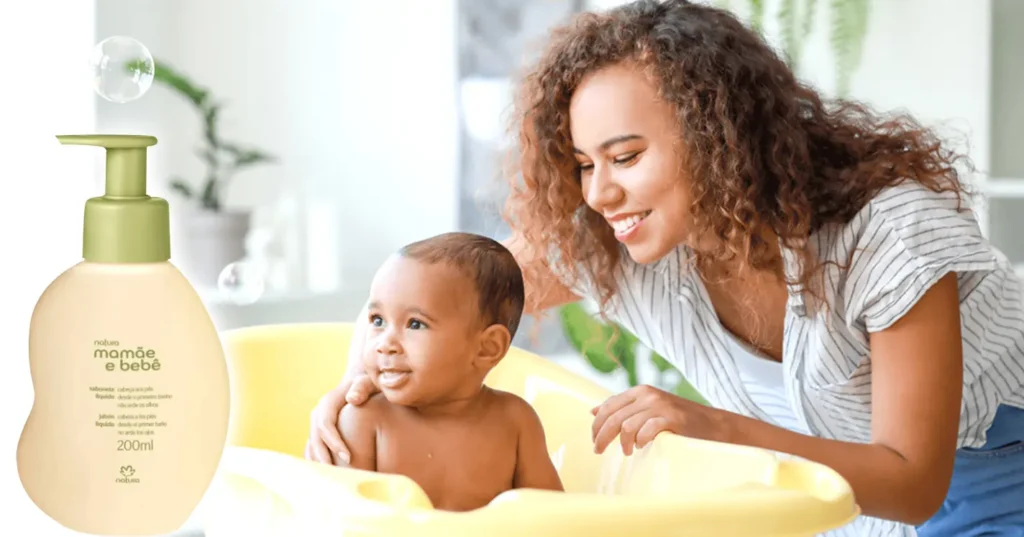 Mãe sorrindo e dando banho em um bebê na banheira amarela, fundo da imagem em leve desfoque com o frasco do Sabonete Líquido da Cabeça aos Pés Natura Mamãe e Bebê em destaque ao lado.